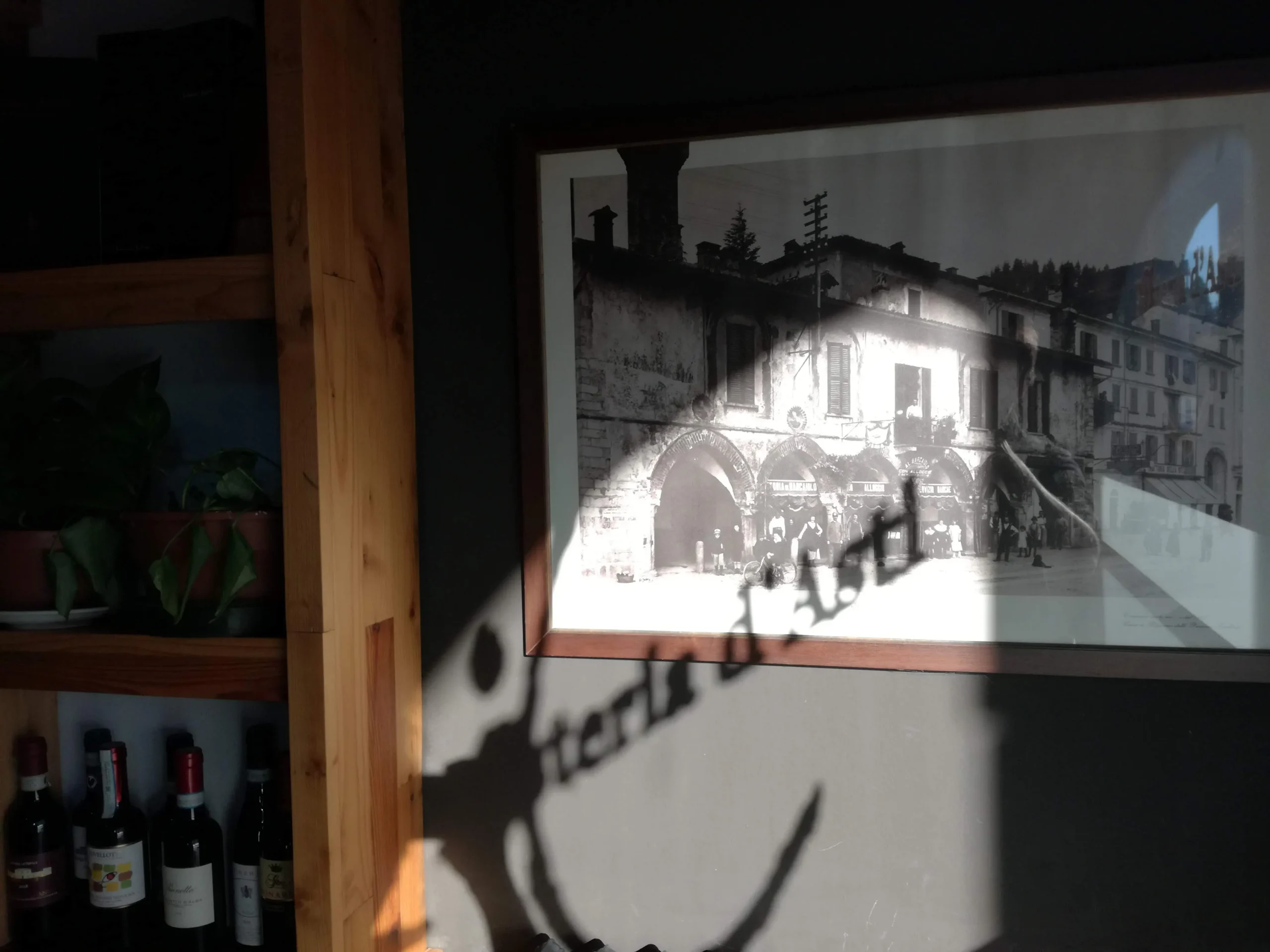 dettaglio dell'interno, con una foto in bianco e nero, l'ombra proiettata sul muro dalla scritta osteria d'asti e delle bottiglie di vino su un mobile in legno